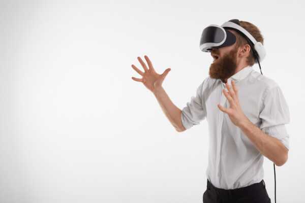 Horizontal image of bearded Caucasian businessman screaming and gesturing emotionally while playing video games using 3d VR goggles, posing at white studio wall with copy space for your text